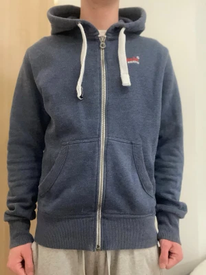 Mörkblå hoodie från Superdry - Mörkblå zip-hoodie från Superdry med vit dragsko och broderad logga på bröstet. Mjukt material, två fickor fram och ribbade muddar. Perfekt för en avslappnad stil.