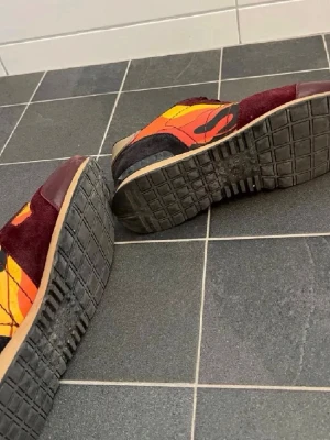 Valentino sneakers i vinrött och orange - Färgstarka sneakers från Valentino med vinröda mockadetaljer, orange och gula paneler samt svarta och beige inslag. Snygg kontrast mellan material och färger, med klassisk låg siluett och robust gummisula. Perfekta för dig som vill sticka ut med din stil.