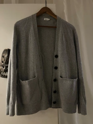 100% Ull kofta från Filippa K - Stilren ull kofta från Filippa K med knäppning framtill och stora fickor. Koftan har långa ärmar, ribbade muddar och en mjuk, tunn stickning som passar perfekt till lager på lager. Enkel och tidlös design som funkar till det mesta.