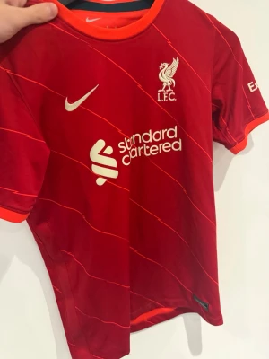 Liverpool matchtröja Nike röd - Säljer en röd Liverpool FC matchtröja från Nike med orange detaljer och tunna diagonala linjer. Tröjan har klubbmärke och sponsortryck framtill samt korta ärmar. Perfekt för fotbollsträning eller att bära på matchdagar. Tröjan är äkta!