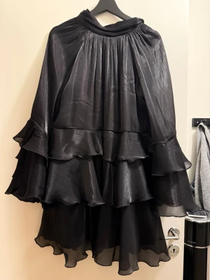 Svart volangklänning med rosett - Supersnygg svart kortklänning med flera lager volanger och en stor rosett. Klänningen  har vida ärmar och ett glansigt material som ger en lyxig känsla. Perfekt för dig som vill sticka ut med en unik och trendig look. Aldrig använd - utan prislapp