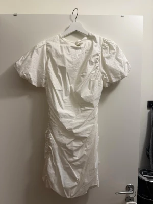 Vit kort puffärmad klänning med knyt - Säljer en vit kort klänning med puffärmar och snygga knytband i ryggen. Klänningen har en öppen rygg med knytband och rynkade detaljer längs sidorna. Endast använd 1 gång