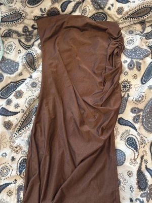 Brun axelbandslös långklänning - Snygg brun långklänning med axelbandslös design och draperad detalj på ena sidan. Perfekt för dig som vill ha en stilren och trendig look. Klänningen har en smickrande passform och är enkel att styla med accessoarer.