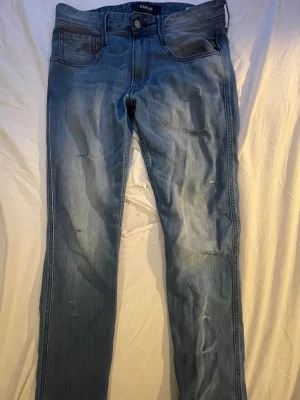 Blå slitna jeans från Replay - Säljer ett par blå jeans från Replay med snygga slitningar och tvättad look. Jeansen har klassisk femficksmodell, raka ben och detaljerad bakficka med Replay-lapp i läder. Perfekta för en avslappnad och trendig stil.