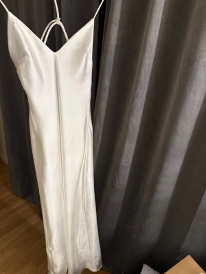 Vit långklänning i satin från Zara - Elegant vit långklänning från Zara i glansigt satinmaterial. Klänningen har smala axelband, v-ringad hals och öppen rygg med snygga banddetaljer. Perfekt för sommarens alla tillfällen.