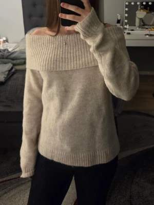 Beige off shoulder stickad tröja från Gina Tricot - Mysig beige stickad tröja med bred ribbad off shoulder-krage och långa ärmar. Tröjan har ribbade muddar vid ärmslut och nederkant, vilket ger en snygg och avslappnad look. Perfekt för dig som gillar en trendig och bekväm stil.