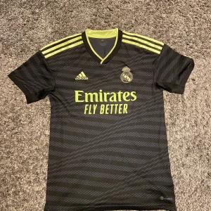 Real Madrid matchtröja svart/gul Adidas - Snygg svart Real Madrid matchtröja från Adidas med gula detaljer och klubbmärke på bröstet. Tröjan har V-ringning, korta ärmar och är tillverkad i ett lätt funktionsmaterial. Perfekt för fotbollsträning eller att bära som supporter.