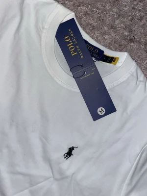 Vit t-shirt från Polo Ralph Lauren - Klassisk vit t-shirt från Polo Ralph Lauren med broderad svart logga på bröstet. T-shirten har rund halsringning och korta ärmar, tillverkad i mjuk bomull för en skön och stilren look.