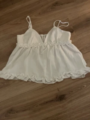 Vit volangig linnetopp - vit linnetopp med tunna axelband och volangdetaljer både upptill och nedtill. Aldrig andvänt 