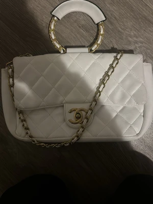 Vit quiltad handväska från Chanel - Snygg vit handväska från Chanel i quiltat läder med guldfärgade detaljer. Väskan har en ikonisk CC-logga på locket, kedjerem i guld och vitt samt ett runt handtag med gulddetaljer. Perfekt accessoar för dig som vill sticka ut med lyxig stil.