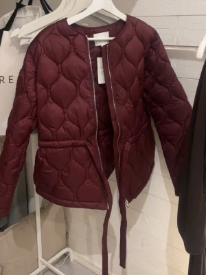 Trendiga Vinröd jacka med bälte - Snygg vinröd vindjacka med quiltat mönster och rund halsringning. Jackan har dragkedja framtill och ett matchande bälte i midjan så formar välldigt fint .