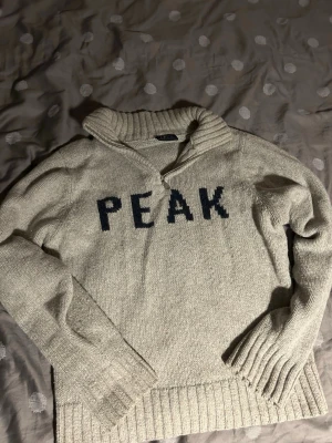 Beige stickad tröja Peak Performance - Beige stickad tröja från Peak Performance med mörkblå text 'PEAK' framtill. Tröjan har hög krage med dragkedja och breda ribbade muddar vid ärmslut och nederkant. Perfekt för kalla dagar och enkel att matcha.