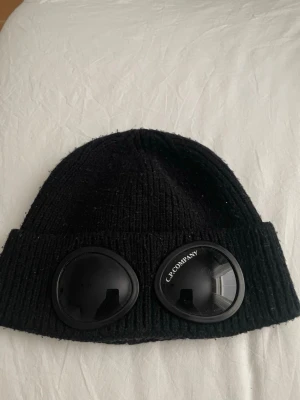 Svart Goggle-mössa från CP - Säljer en svart ribbstickad mössa från Cp  med de ikoniska goggle-linserna framtill. Mössan är i mjukt material och har en tight passform som sitter snyggt på huvudet. Perfekt för dig som vill ha en streetwear-inspirerad look.