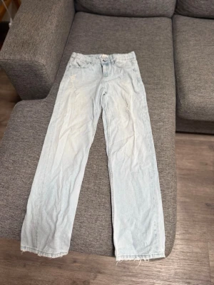 Ljusblå raka jeans med rå kant - Säljer ett par ljusblå jeans med rak passform och råa, fransiga benslut. Jeansen har diskreta slitningar framtill och klassiska fem fickor. De är från Gina Tricot young i storlek 152