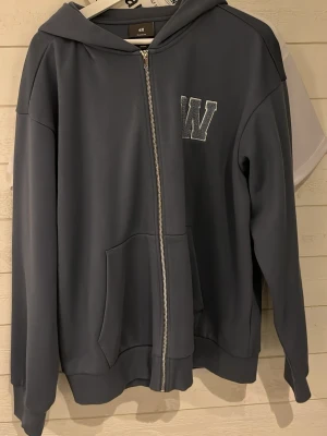 Mörkgrå hoodie med dragkedja H&M - Mörkgrå hoodie från H&M med dragkedja framtill och broderad W på bröstet. Klassisk modell med huva och känguruficka. Perfekt för en avslappnad stil och enkel att matcha med jeans eller joggers.