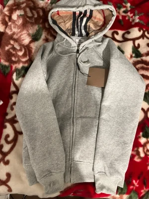 Grå zip hoodie från Burberry - Snygg grå hoodie från Burberry med dragkedja framtill och klassiskt rutigt foder i huvan. Hoodien har ribbade muddar vid ärmslut och nederkant samt dragsko i huvan. Perfekt för en avslappnad men stilren look. Materialet är mjukt och bekvämt, troligen bomullsblandning.