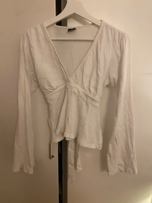 Vit topp från Gina med knytband 🤍 - Snygg vit topp med v-ringning och långa ärmar. Toppen har knytband i midjan som ger en fin passform. Perfekt att styla med jeans eller kjol för en trendig look. 💕 (använd några få gånger)