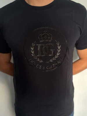 Svart Dolce & Gabbana t-shirt - Snygg svart t-shirt från Dolce & Gabbana med diskret glansigt DG-logotyptryck och krona på bröstet. Klassisk rund halsringning och korta ärmar. Perfekt för dig som vill ha en stilren och exklusiv look.