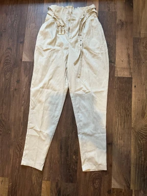 Vita linnebyxor med bälte - Stilrena beiga linnebyxor med hög midja och dubbla bälten i midjan. Byxorna har avsmalnande ben och är perfekta för en avslappnad men trendig look. Materialet är linne vilket ger en luftig känsla.                                     