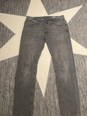Grå raka jeans från Jack&Jones  - Säljer ett par grå jeans från Jack&Jones med rak passform och klassisk femficksdesign. Jeansen har en snygg tvättad look och är tillverkade i denim. Perfekta för dig som gillar en avslappnad stil.