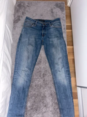Blå raka jeans från Nudie Jeans - Säljer ett par klassiska blå jeans från Nudie Jeans med raka ben och femficksmodell. Jeansen har en snygg tvätt med slitningar på låren och bakfickor med Nudie-logga. Perfekta till vardags och passar till det mesta. | W28 L32 / Finns en gräs fläck på knät, men det syns knappast. Pris kan förhandlas 
