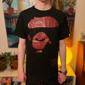 Svart BAPE t-shirt med röd logga - Cool svart t-shirt från BAPE med det ikoniska röda apansikte-trycket på bröstet. Klassisk passform med rund hals och korta ärmar. Perfekt för dig som gillar streetwear och vill sticka ut med en statement-tee.