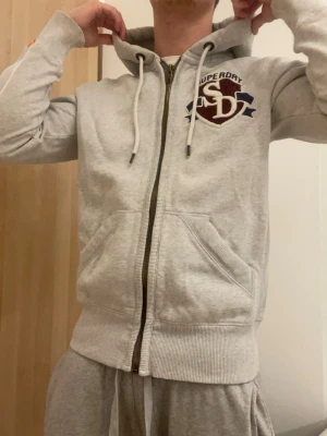Grå hoodie med dragkedja från Superdry - Snygg ljusgrå hoodie från Superdry med dragkedja, huva med vita snören och broderad  logga. ⚠️En liten deffekt på luvan (sista bilden)⚠️