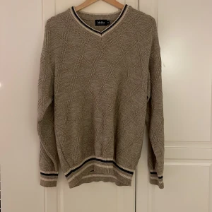 Beige stickad tröja från Melka - Stickad beige tröja från Melka med v-ringad hals och diskret mönster i strukturen. Tröjan är ribb stickad och i storlek M 