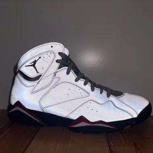 Nike Air Jordan 7 Retro grå/reflective - US size 14. Säljer ett par Nike Air Jordan 7 Retro med grå reflexskinn, svarta och röda detaljer samt guld Jumpman-logga på sidan. Skorna har högt skaft, snörning och svart sula med röda och vita inslag. Insidan är fodrad i rött tyg för extra kontrast. Perfekt för dig som gillar klassiska sneakers med unik design.