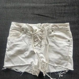 Vita denimshorts med snörning - Säljer ett par vita denimshorts med låg midja och snygg snörning framtill. Shortsen har råa, fransiga kanter och klassiska fickor både fram och bak. Perfekta för sommaren och ger en cool, avslappnad vibe.