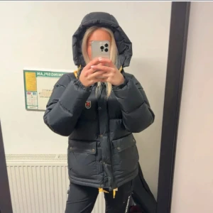 Svart pufferjacka från Fjällräven L - Säljer en svart pufferjacka från Fjällräven i storlek L. Jackan har huva med gula snören, stora fickor med tryckknappar och klassisk Fjällräven-logga på bröstet. Perfekt för kalla dagar med sin dunfyllda, quiltade design och snygga detaljer. Jackan är i mycket bra skick säljer då jag vill köpa en ny jacka till vintern 💕