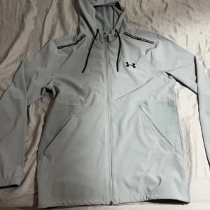 Ljusgrå vindjacka från Under Armour - Snygg ljusgrå vindjacka från Under Armour med huva, svarta snören och dragkedja framtill. Jackan har två stora fickor, perforerade detaljer på sidorna och logga på bröstet. Perfekt för dig som gillar sportig och clean stil.