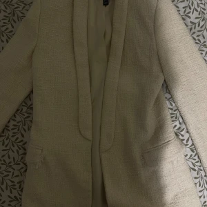 Beige kavaj från Mango i tweed - Säljer en beige kavaj från Mango i tweed-look med råa kanter vid fickorna. Kavajen har en klassisk krage, lång ärm och är helfodrad. Perfekt för dig som vill ha en stilren och trendig look.