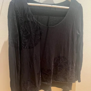 Svart långärmad blus med sammet - Svart blus från Desigual i storlek S med lång ärm och rund halsringning. Blusen har dekorativa sammetspartier med mönster i form av cirklar och blad på både ärmar och nedtill. Materialet är mjukt och har en lätt glansig finish som ger en cool look.