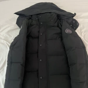 Svart dunjacka från Canada Goose XS - Säljer en svart dunjacka från Canada Goose i storlek XS. Jackan har huva, knappar framtill och klassisk patch på ärmen. Perfekt för kalla dagar med sin tjocka och vadderade design. Snygg och stilren modell som håller dig varm hela vintern.