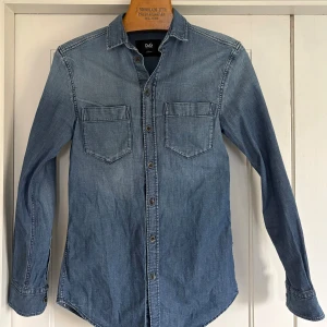 Blå jeansskjorta, Dolce & Gabbana - Säljer en snygg blå jeansskjorta från Dolce & Gabbana med två bröstfickor. Skjortan har klassisk krage, lång ärm och är i slim fit-modell. Perfekt för dig som gillar stilren denim och vill ha en vintage skjorta!