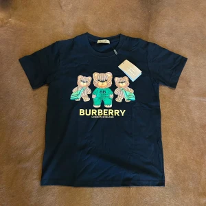 Svart Burberry t-shirt med nalletryck - Svart t-shirt från Burberry med coolt tryck av tre stickade nallar på bröstet och tydlig Burberry-logga i gult. Klassisk rund halsringning och korta ärmar. Materialet är mjuk bomull som känns skönt mot huden. Perfekt för dig som vill sticka ut med en lekfull designerstil.