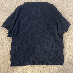 Svart kortärmad blus med struktur - Säljer en svart blus med korta ärmar och en cool, strukturerad yta. Toppen har en loose passform och är tillverkad i ett mjukt, syntetiskt material som ger en skön känsla. Perfekt för dig som gillar minimalistisk stil med lite edge. Tröjan har en lite högre krage. 