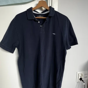 Mörkblå pikétröja Hampton Republic - Klassisk mörkblå pikétröja från Hampton Republic med vit broderad logga på bröstet. Tröjan har krage, knäppning med tre knappar och korta ärmar. Tillverkad i bomull för en skön och luftig känsla. Perfekt för en stilren och avslappnad look.