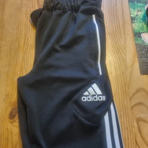 Svarta Adidas mjukisbyxor med vita ränder - Svarta mjukisbyxor från Adidas med klassiska vita ränder längs sidorna och broderad logga på benet. Byxorna har elastisk midja och är gjorda i mjukt material som är perfekt för chill eller träning. Snygg och sportig look.