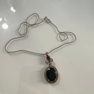 Halsband - Unikt halsband med tunn silverfärgad kedja och stort ovalt hänge. Hänget har en mörkgrön slipad sten i mitten, omgiven av små klara stenar och detaljer i guldton. Ovanför sitter två klara stenar och en mindre rosa sten. Kedjan är 42cm lång