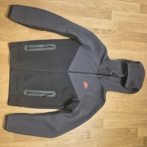 Svart Nike hoodiejacka med dragkedja XS - Nike hoodiejacka i svart med mörkgrå detaljer och röd logga på bröstet. Jackan har hel dragkedja, två dragkedjeförsedda fickor framtill och huva. Tillverkad i mjukt material, perfekt för en sportig och avslappnad stil.
