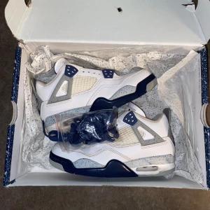 Nike Air Jordan 4 vit/blå sneakers - Nike Air Jordan 4 sneakers i vitt och marinblått med grå och blå stänkdetaljer på häl och sula. Skorna har snörning, platt sula och rund tå. Ikonisk Jumpman-logga i blått och rött på häl och undersida. Tillverkade i skinn och syntetmaterial. Levereras med originalkartong och extra skosnören.