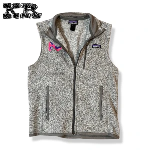 Grå fleeceväst från Patagonia - Gråmelerad fleeceväst från Patagonia med hel dragkedja framtill och två sidofickor med dragkedja. Västen har en hög krage och ett broderat märke på bröstet i lila och rosa. Perfekt för lager-på-lager och chill stil.