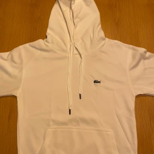 Vit hoodie från Lacoste med huva - Vit hoodie från Lacoste med klassisk krokodillogga på bröstet. Tröjan har huva med snörning, stor magficka och är tillverkad i 100% bomull. Perfekt för dig som gillar cleana och stilrena plagg med sportig vibe.