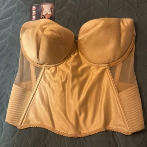 Guldig korsett med mesh och bred resår - Snygg guldig korsett med vadderade kupor och breda ribbade resårpartier i sidorna. Toppen har meshdetaljer och stängs med hyskor i ryggen. Materialet är glansigt och ger en lyxig känsla. Perfekt för att skapa en statement-look.