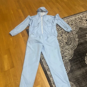 Ljusblå vindjacka från Lacoste - Säljer en ljusblå vindjacka från Lacoste med huva och snörning. Jackan har en stor krokodil-illustration på ryggen och Lacoste-logga på ena ärmen. Dragkedja framtill och elastiska muddar. Perfekt för dig som gillar streetwear och vill sticka ut.