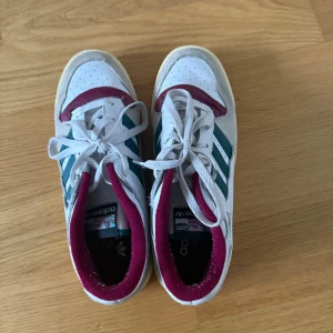 Adidas sneakers med hög sula  - Snygga Adidas sneakers med klassiska tre ränder i teal på sidan, vit ovandel i skinn och mocka, samt vinröda detaljer runt kragen och tån. Platt sula i beige och vit, snörning framtill och rund tå. Perfekt för dig som gillar retrostil och färgkombinationer.