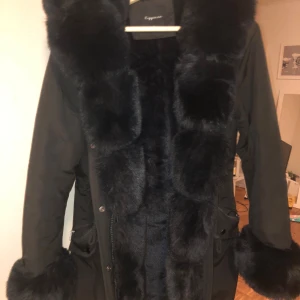 Svart parkas med fuskpäls  - Säljer en svart parkas med stora fluffiga fuskpälsdetaljer längs krage, ärmslut och insida. Jackan har dragkedja, snörning i midjan och praktiska fickor. Perfekt för kalla dagar när du vill vara både varm och snygg, jag har bara använt den några gånger den är i storlek L men den är liten i modellen så den passa bra för dig som ha storlek M eller S den har mjuk päls insidan och är väldig varm att ha på sig, jackan är i mycket fint skick, inga skador eller fläckaroch alla knappar finns kvar.