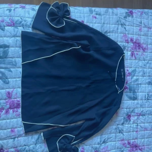 Mörkblå blus med volangärmar - Snygg mörkblå blus från Zara med vita kontrastkanter och unika volangärmar med rosettdetalj. Blusen har en lös passform och tunna axelband som ger en cool offshoulder-look. Perfekt för dig som vill sticka ut med stilrena detaljer.